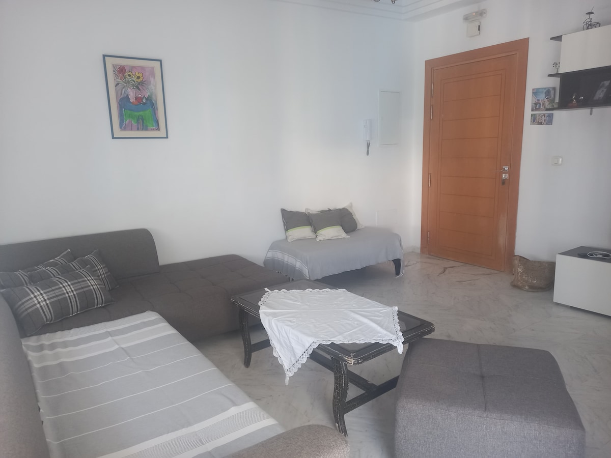 An inviting living area features a spacious gray sofa and a plush ottoman. A small coffee table sits in the center, accented by a light tablecloth. A light-colored bed is positioned nearby, and artwork is displayed on a white wall, enhancing the room's warmth.