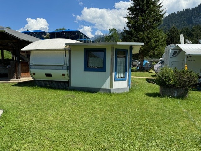 A caravan with an attached awning is situated on a grassy area. The exterior features a combination of blue and white, enhancing its inviting appearance. Surrounding greenery contributes to a calm outdoor space, with mountains visible in the background under a partly cloudy sky.