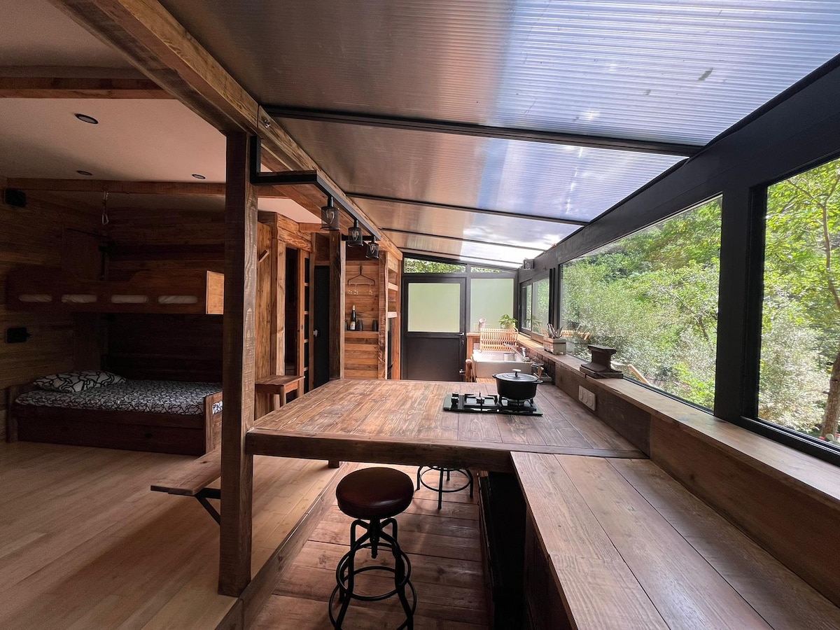 An open kitchen area features a wooden dining table with a built-in cooking stove. Natural light floods through expansive windows that provide a view of the surrounding greenery. The adjoining sleeping area includes a cozy bed with patterned bedding, complementing the rustic wooden interior.