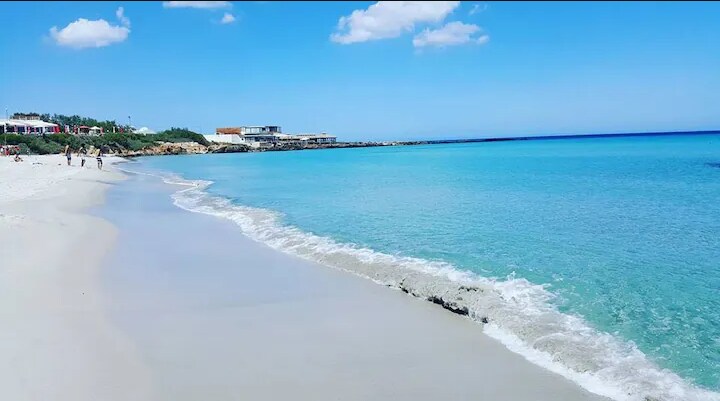 A serene beach scene features soft, white sand gently meeting calm, turquoise waters. Soft waves create a rhythmic shoreline, while a clear blue sky is dotted with a few fluffy clouds. In the distance, beachside establishments can be seen along the sandy stretch.