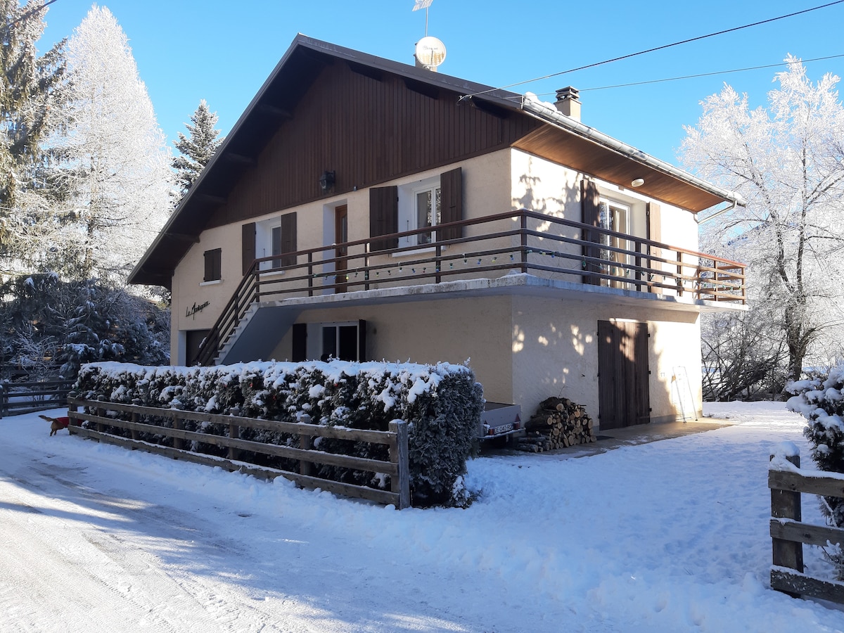 A chalet-style building is featured, set in a snowy environment. The lower level has a welcoming concrete entrance, while the upper balcony is accessible by external steps. Surrounding greenery is adorned with frost, and a wooden fence lines the front, framing the entrance.