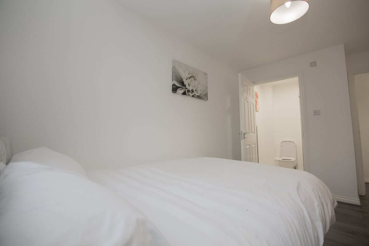 A bright and minimalist bedroom is presented, featuring a spacious bed with white bedding. A subtle floral artwork is displayed on the wall. A doorway leads to a small bathroom area, which is visible in the background.