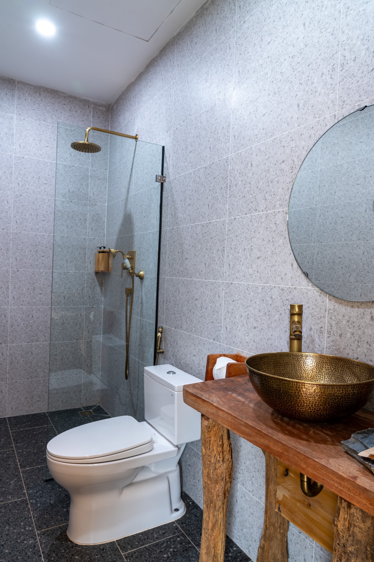 A modern bathroom features a walk-in shower enclosed with clear glass, showcasing a sleek rainfall showerhead. A round mirror is mounted above a stylish wooden sink with bronze detailing. The area is complemented by textured grey tiles and natural wood accents, highlighting a clean and organized space.