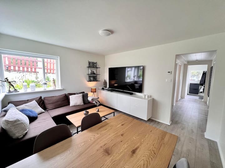Luminosa sala de estar de planta abierta centrada en una elegante mesa de comedor de madera y un gran y cómodo sofá seccional. Disfruta del entretenimiento a través del enorme televisor inteligente montado en la pared y la barra de sonido, todo apoyado por elegantes gabinetes flotantes blancos.