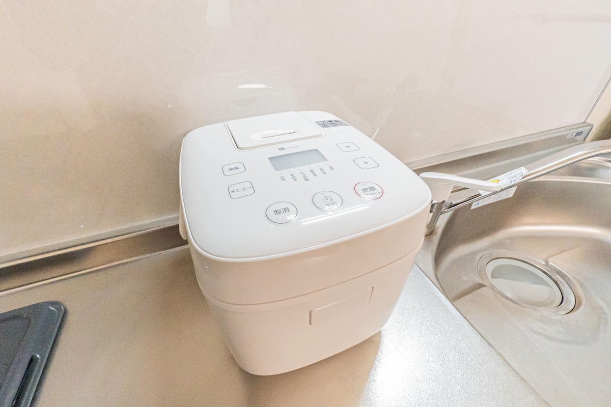 A rice cooker is placed on a kitchen counter. The appliance features a digital display and various buttons, designed for easy operation. The counter is made of a smooth material, providing a clean backdrop for the cooker.