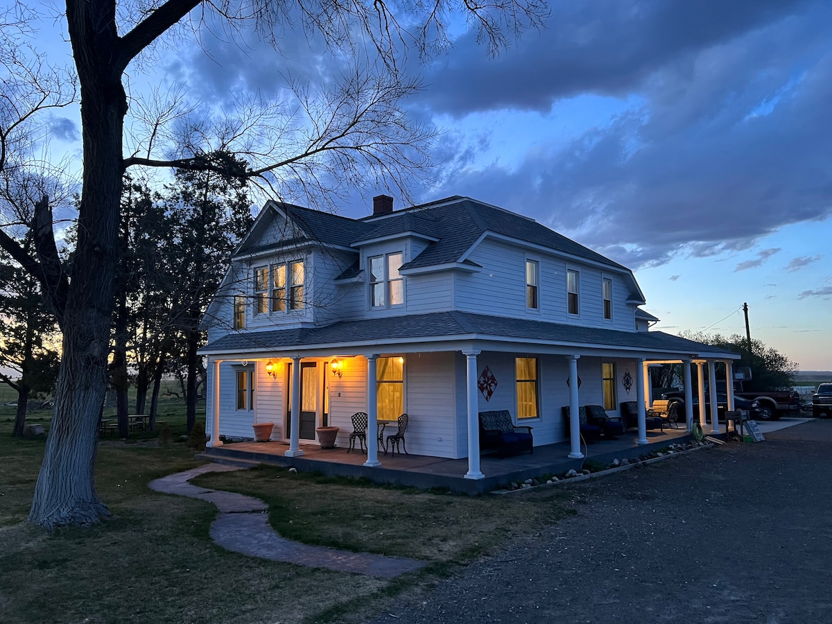 The exterior of the historic Hudson House Bed and Breakfast is portrayed at dusk, showcasing a two-story structure with multiple windows and inviting outdoor seating. Soft lighting emanates from within, illuminating the porch and creating a welcoming atmosphere.