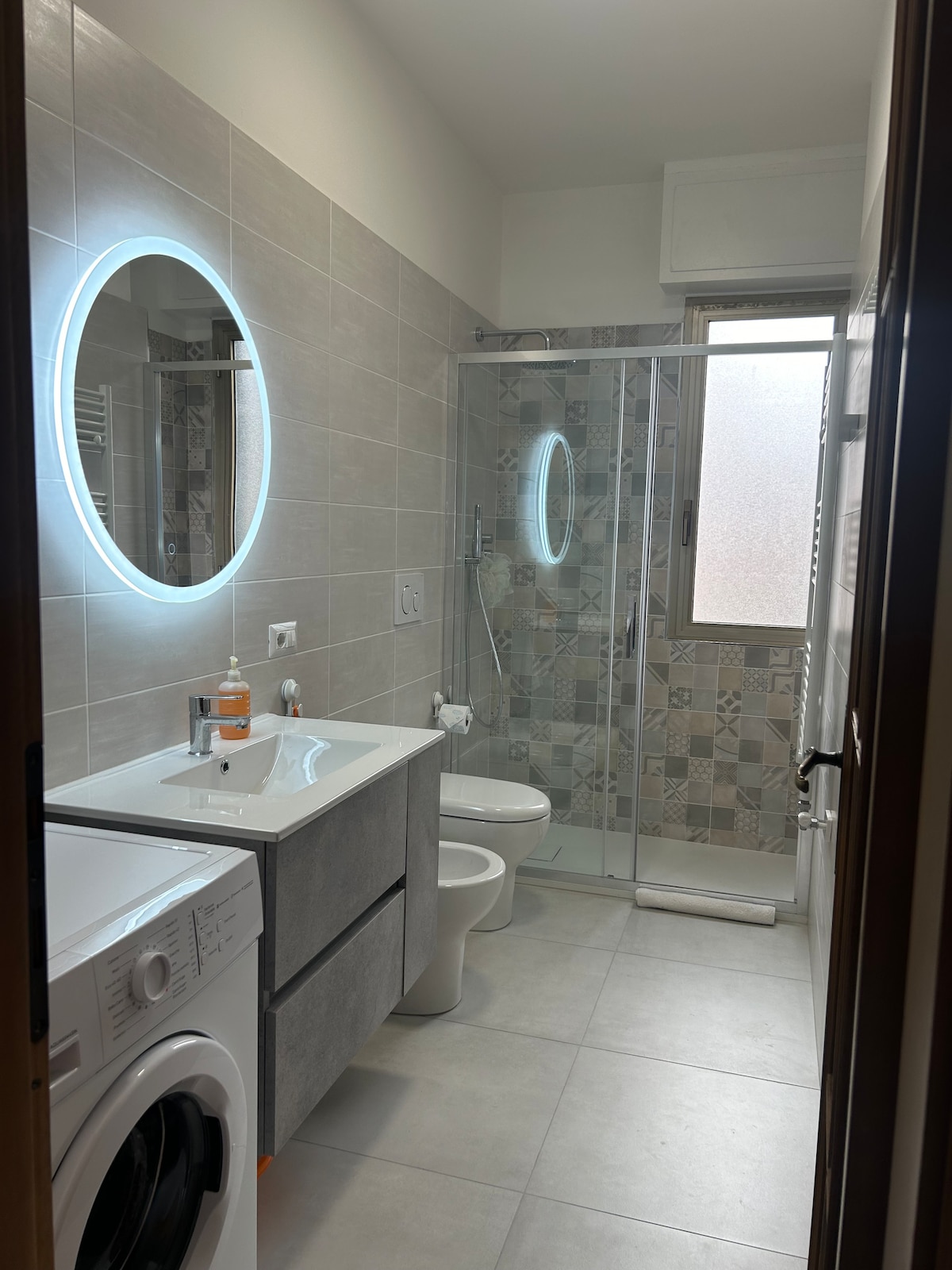 The bathroom features modern fixtures including a large shower enclosed by glass. A round mirror with LED lighting is mounted above the sink. The space includes a washing machine and a toilet, with tiled walls that incorporate a patterned design.