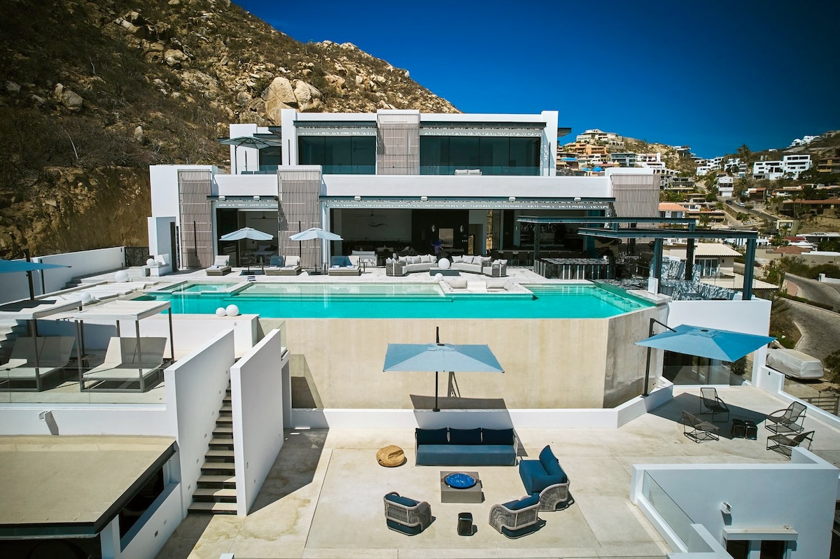 An aerial view captures a sprawling villa with a large infinity pool at its center, surrounded by spacious deck areas featuring lounge chairs and umbrellas. The modern architecture is highlighted against a clear blue sky, with scenic hillside homes visible in the background.