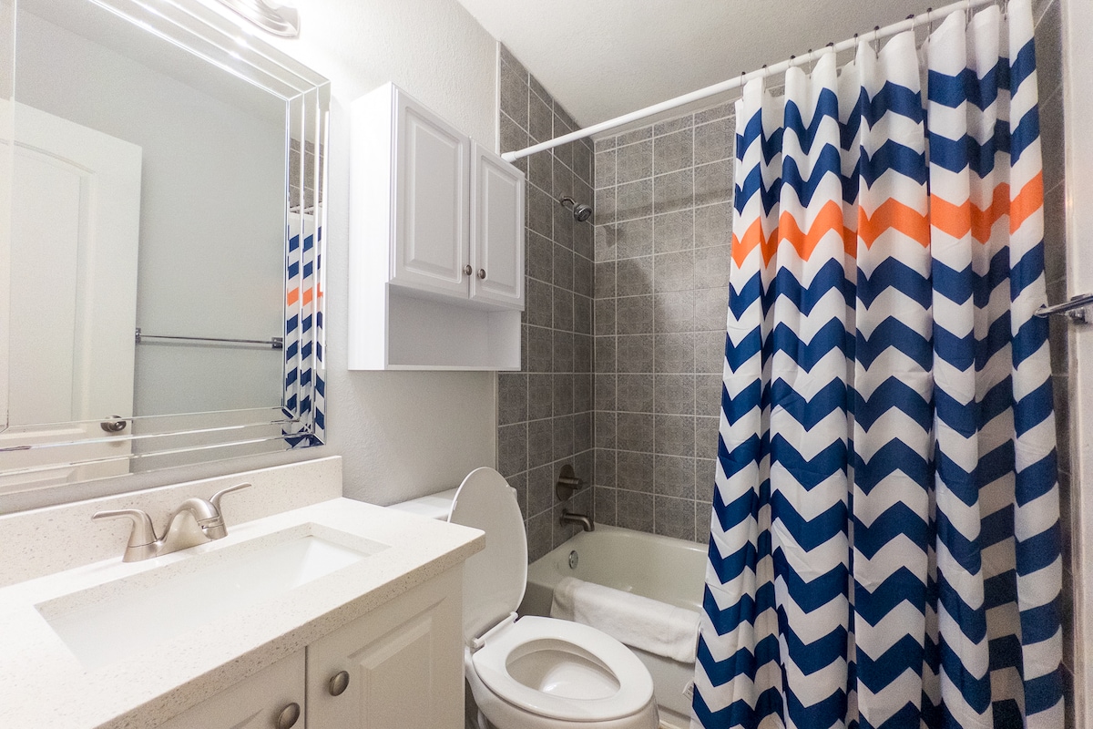 The bathroom features a modern design with a white countertop and mirror. A bathtub is equipped with a shower, which is adorned with a blue and orange chevron-patterned curtain. Storage is provided by wall-mounted shelves, and the tiled wall complements the overall aesthetic.