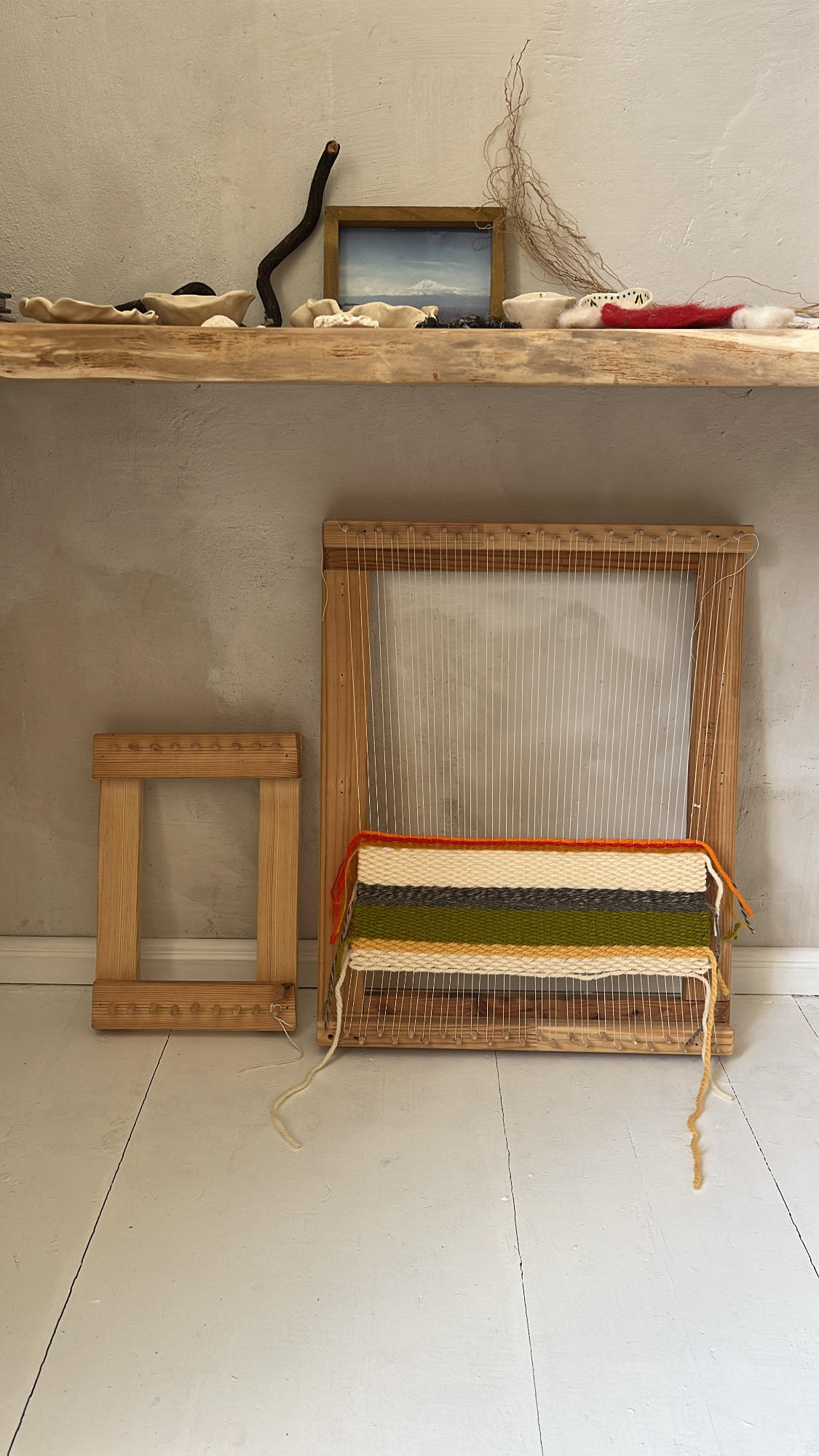 A wooden weaving loom stands beside a matching frame. Various crafting materials are displayed on a shelf above, including natural elements and small artwork, contributing to a creative atmosphere.