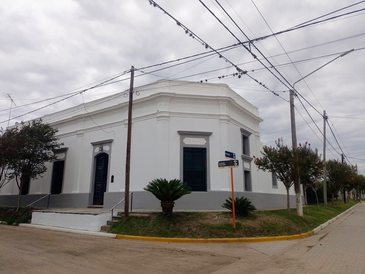 An exterior view of a colonial-style house with a white facade and contrasting dark trim. Large windows framed in black are visible, and the structure is surrounded by well-maintained landscaping, including palm trees. Power lines run above, and a signpost is positioned nearby.