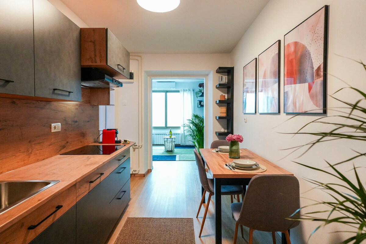A modern kitchen is shown with dark cabinetry and wooden countertops. A dining table is set for two, adorned with a flower centerpiece. Natural light enters through a window at the far end, illuminating the open layout that connects to a living area.