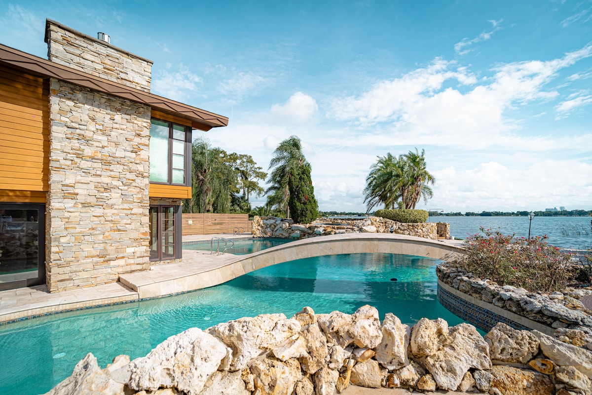 A modern lakeside resort is showcased, featuring a stone exterior and a sweeping pool that curves gently around the patio. Lush palm trees and vibrant landscaping frame the water view, offering a tranquil setting for relaxation.