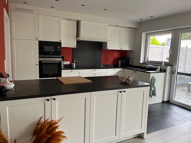 A modern kitchen features sleek white cabinetry and dark countertops. Built-in appliances are integrated into the cabinetry, and a large window provides natural light, with a view of the outdoor area. A wooden cutting board is placed on the central island, enhancing functionality.