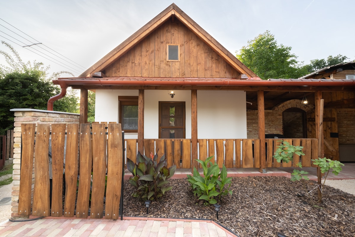 The exterior of a wooden house is seen with a sloped roof and a warm brown facade. A fenced area made of wooden planks surrounds the entrance, which features a door with a small window. Plant beds with greenery are visible in the foreground.