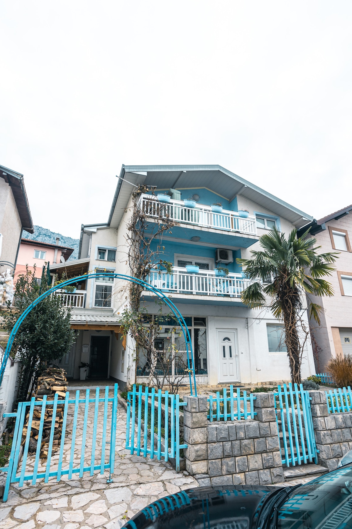 A three-story building with a light blue façade and white trim is depicted. A palm tree and shrubs are visible in the front yard. The entrance is framed by a decorative archway, and a stone wall with a blue picket fence outlines the property.