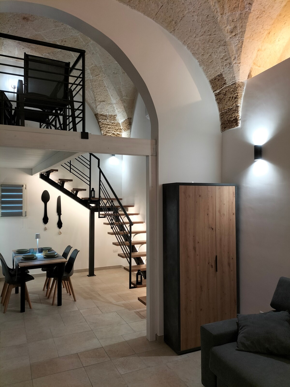 An open-concept living area features high vaulted ceilings with stone arches, complemented by warm lighting. A dining table set for four sits adjacent to a modern kitchenette. An elegant staircase leads to an upper level, showcasing minimalist furniture and a neutral color palette.