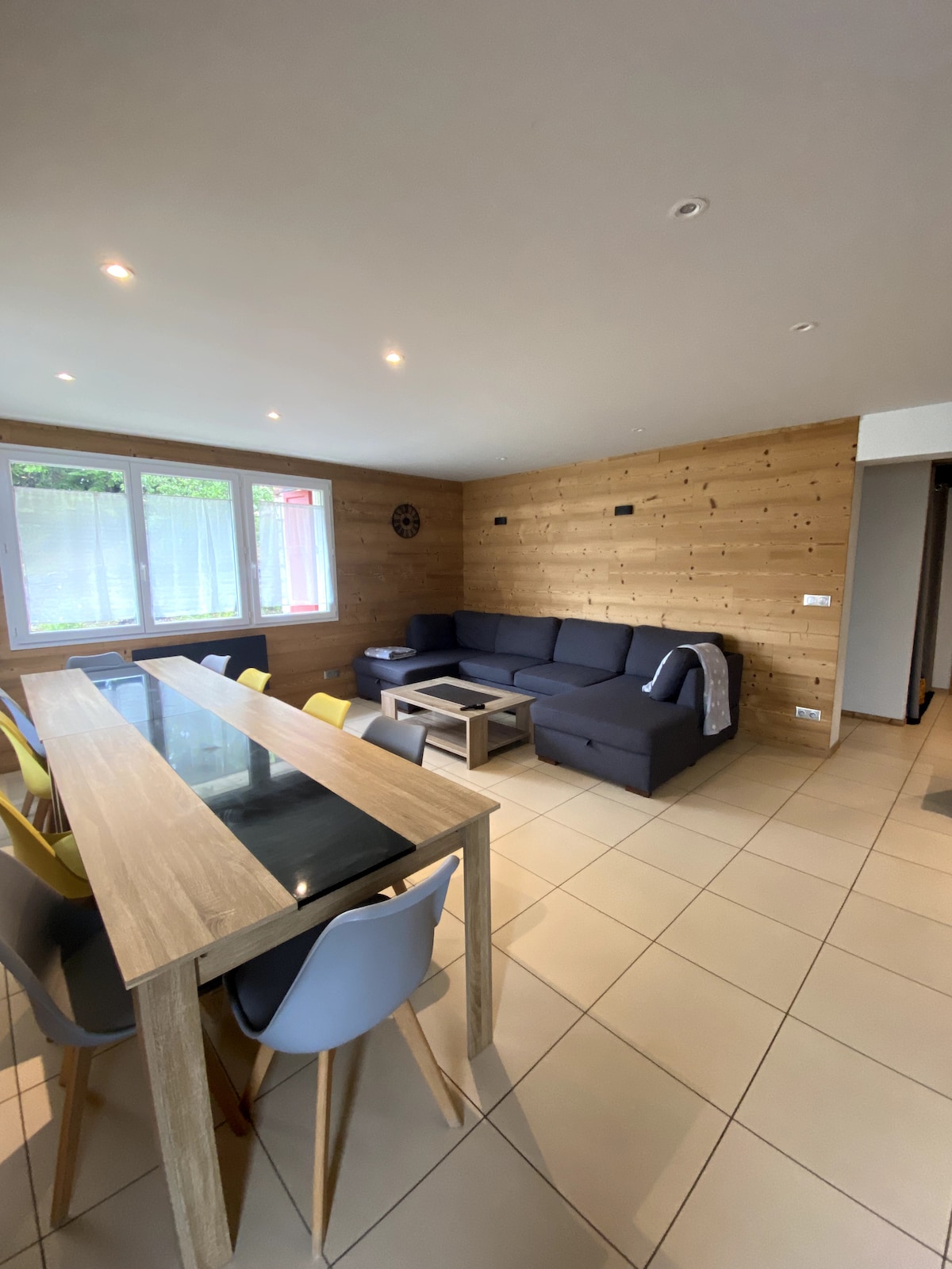 An open-plan living area features a dark sofa arranged around a coffee table, complemented by a dining table with colorful chairs. Large windows allow natural light to fill the space, highlighting the wooden accent walls and tiled floor.