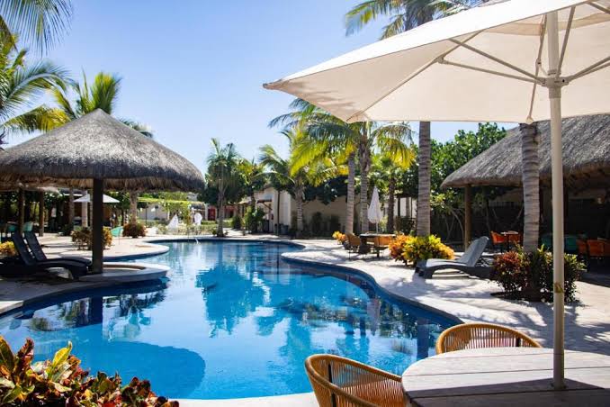 A clear, inviting pool is surrounded by palm trees and tropical foliage. Thatched-roof cabanas provide shaded seating areas, and lounge chairs are situated around the pool's edge. Sunlight reflects off the water, contributing to a serene outdoor environment.