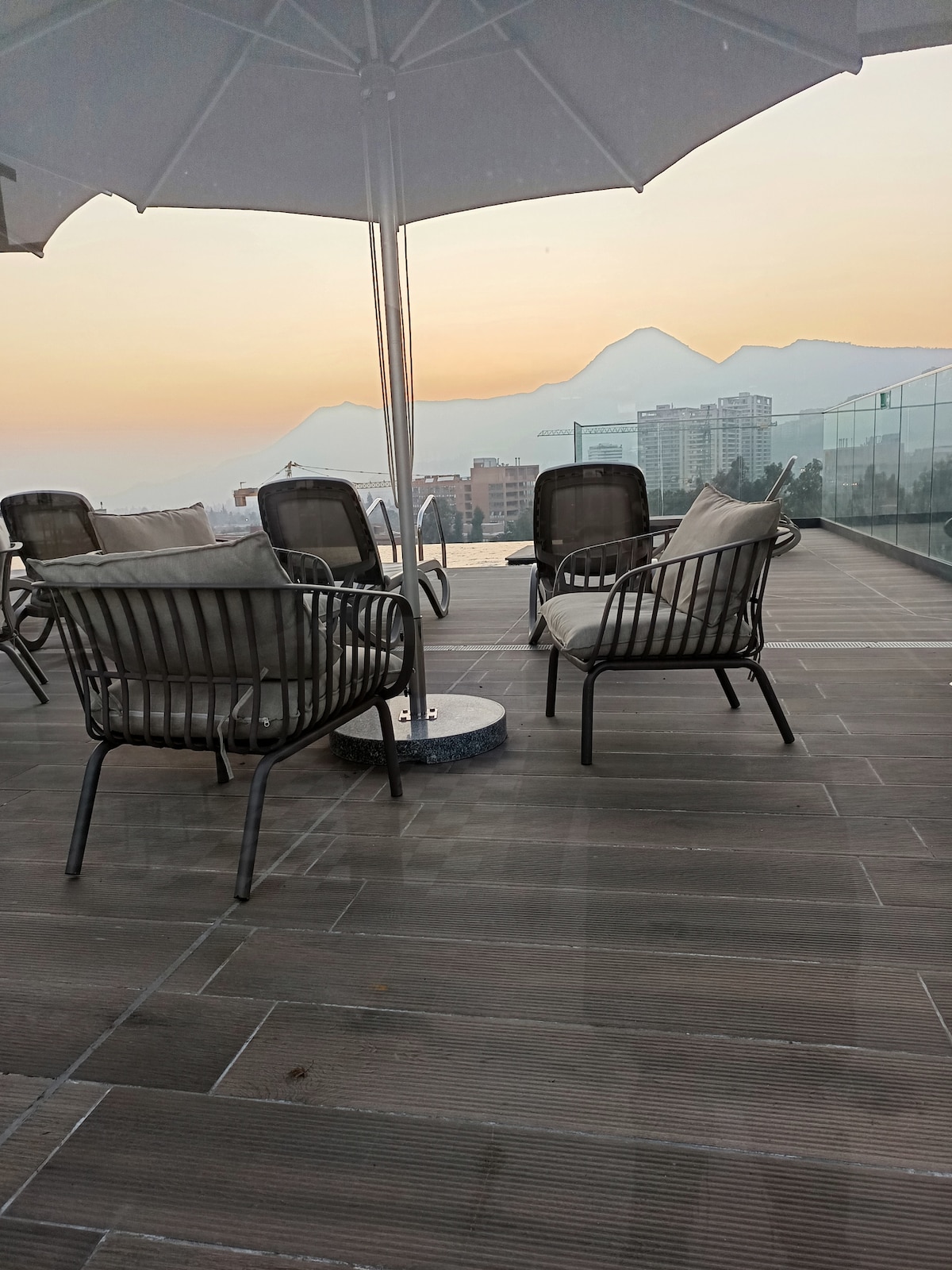 A serene rooftop terrace is presented, featuring several metal lounge chairs with comfortable cushions. An umbrella provides shade, while a distant mountain range is visible under a soft sunrise sky. The expansive view encompasses surrounding buildings, creating a peaceful outdoor gathering space.