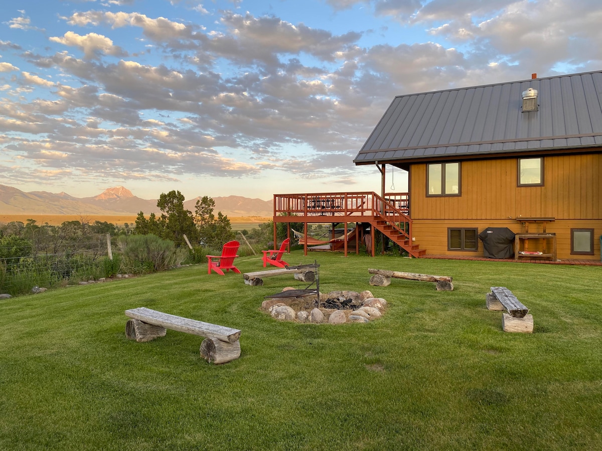A grassy outdoor area features a fire pit surrounded by seating logs, offering a gathering space under a spacious sky. Nearby, two bright red chairs sit invitingly on the lawn, with wooden stairs leading up to the deck of the home in the background.