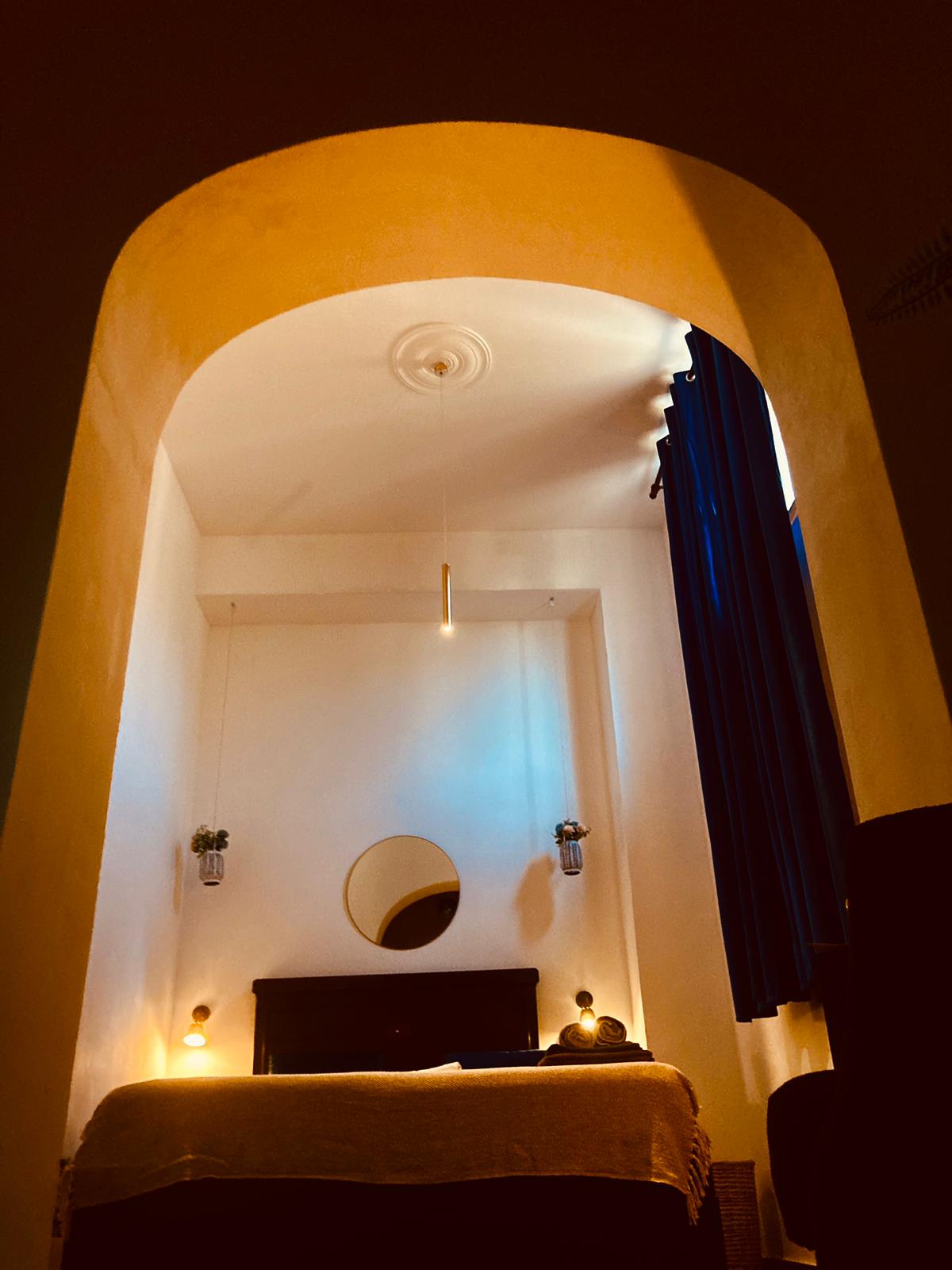 An archway leads to a cozy sleeping area, illuminated by soft light. A circular mirror hangs above a dark wooden headboard, flanked by bedside lamps. Deep blue curtains frame a window, providing a contrast to the light walls and creating a serene environment.