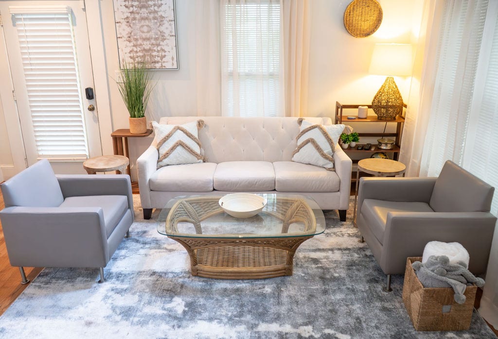 A bright living space showcases a light fabric sofa with tufted detailing, accompanied by two sleek armchairs in a subtle gray. A glass coffee table sits in the center, resting on a soft area rug. Natural light filters through sheer curtains, enhancing the room's openness.