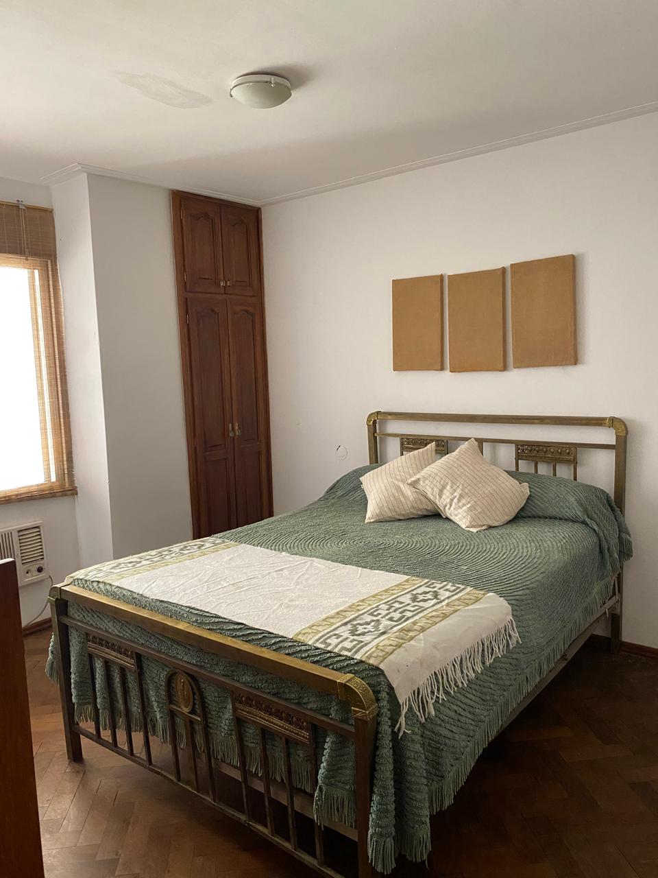 A cozy bedroom is furnished with a spacious bed, complemented by decorative pillows. Natural light enters through a window with wooden blinds. A wardrobe is positioned against the wall, enhancing the room's functionality with minimal decor.