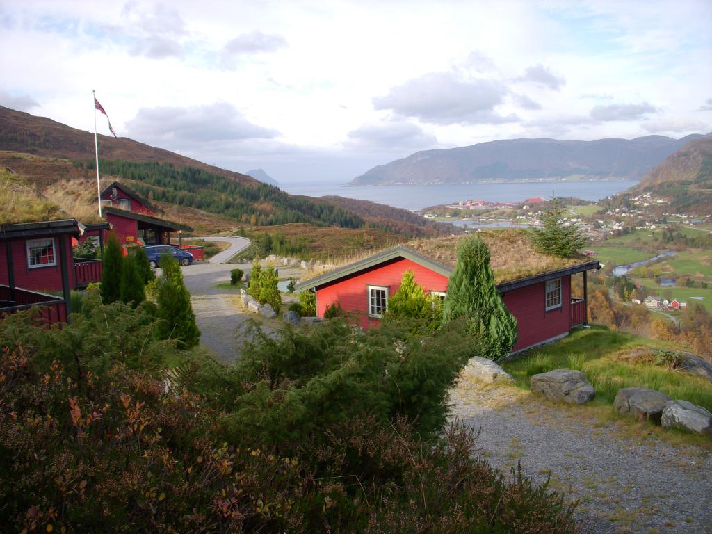 A scenic view features traditional red cabins nestled among greenery, with a winding gravel path leading through the landscape. Mountains and a fjord are visible in the background, complementing the serene natural surroundings.
