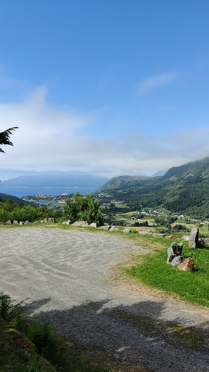 A scenic view from the property reveals sprawling landscapes of green hills and distant mountains, with a fjord visible in the background. The foreground features a gravel area surrounded by grass and stones, highlighting the natural beauty of the location.