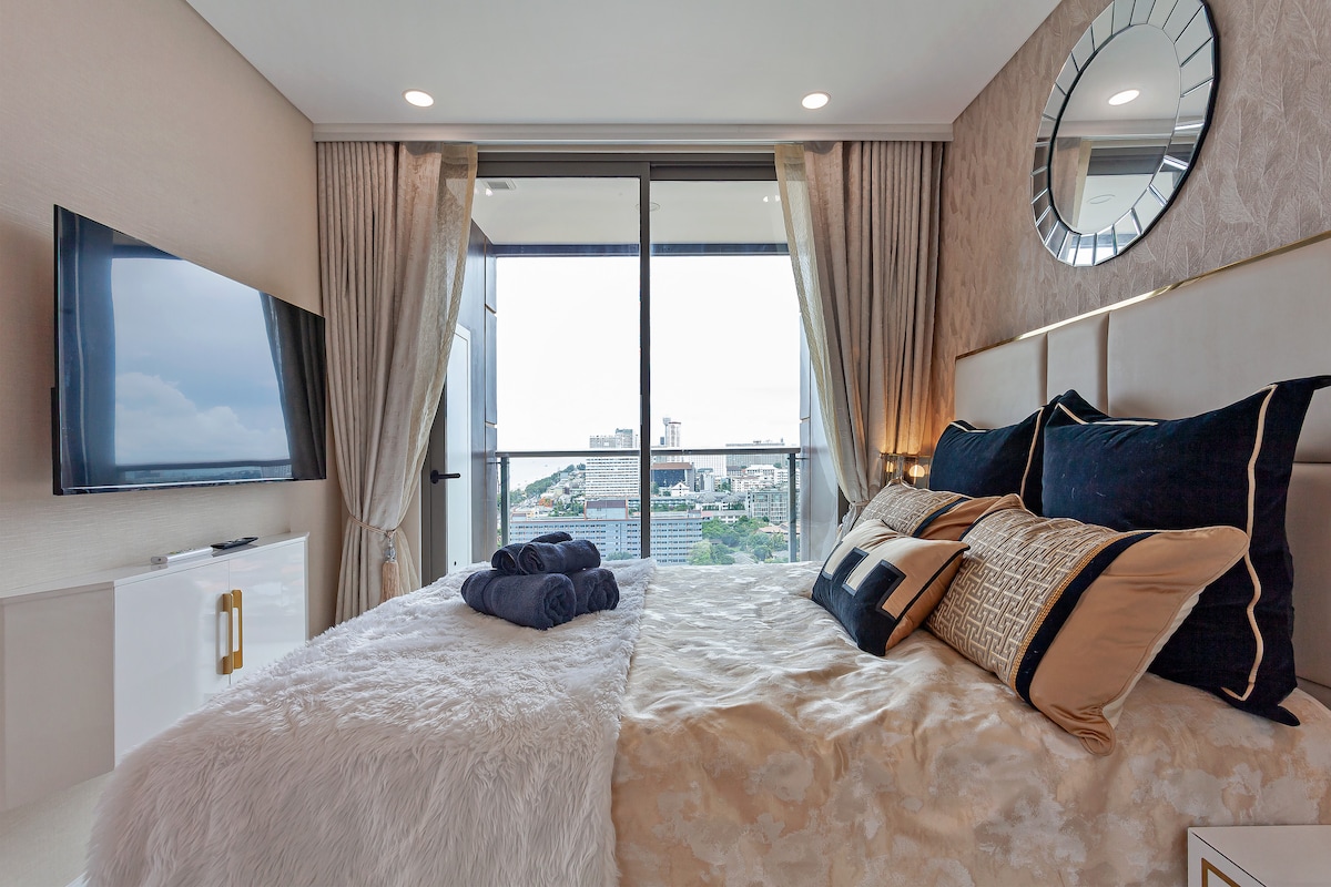 A well-appointed bedroom features a spacious bed with decorative pillows. A large window provides natural light and city views, while a flat-screen TV is mounted on the opposite wall. Elegant curtains frame the window, complementing the overall neutral color palette.