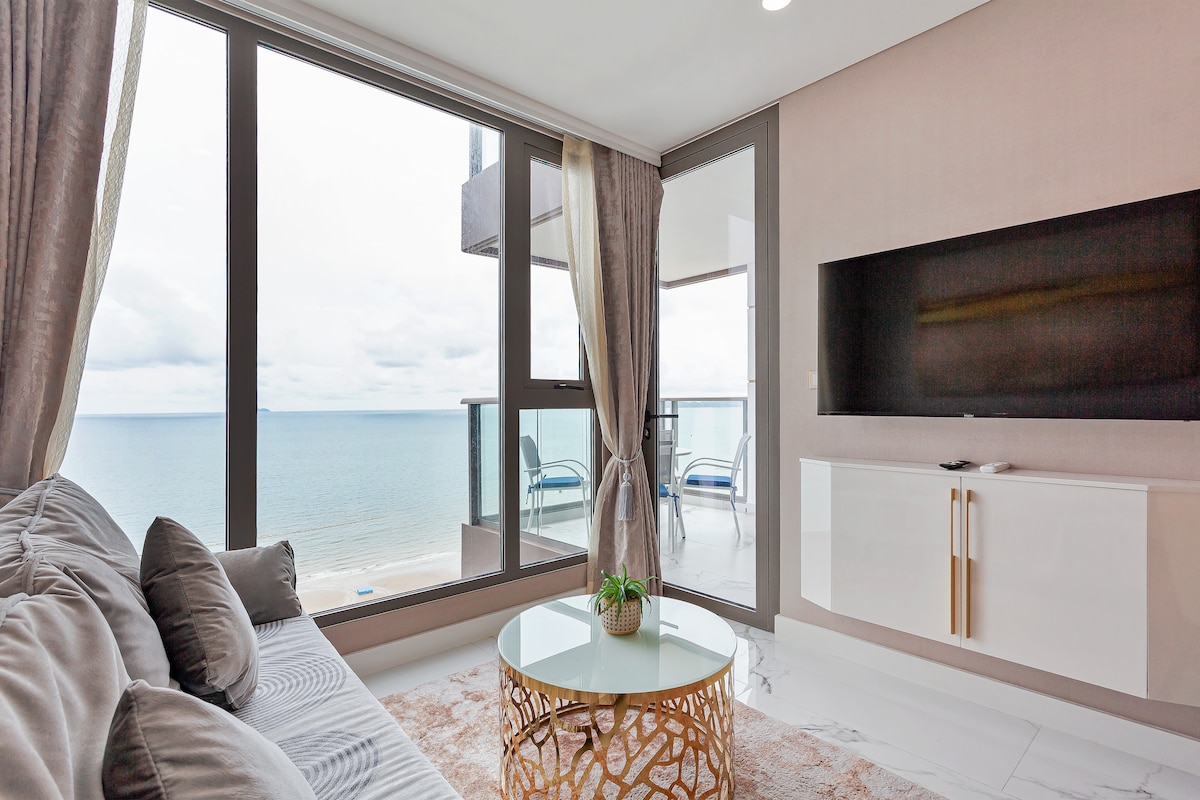 A modern living area features a comfortable sofa with decorative cushions, facing large windows that overlook the beach and ocean. A round coffee table is positioned centrally, while a flat-screen TV is mounted on the wall. Natural light fills the space, enhancing the airy ambiance.