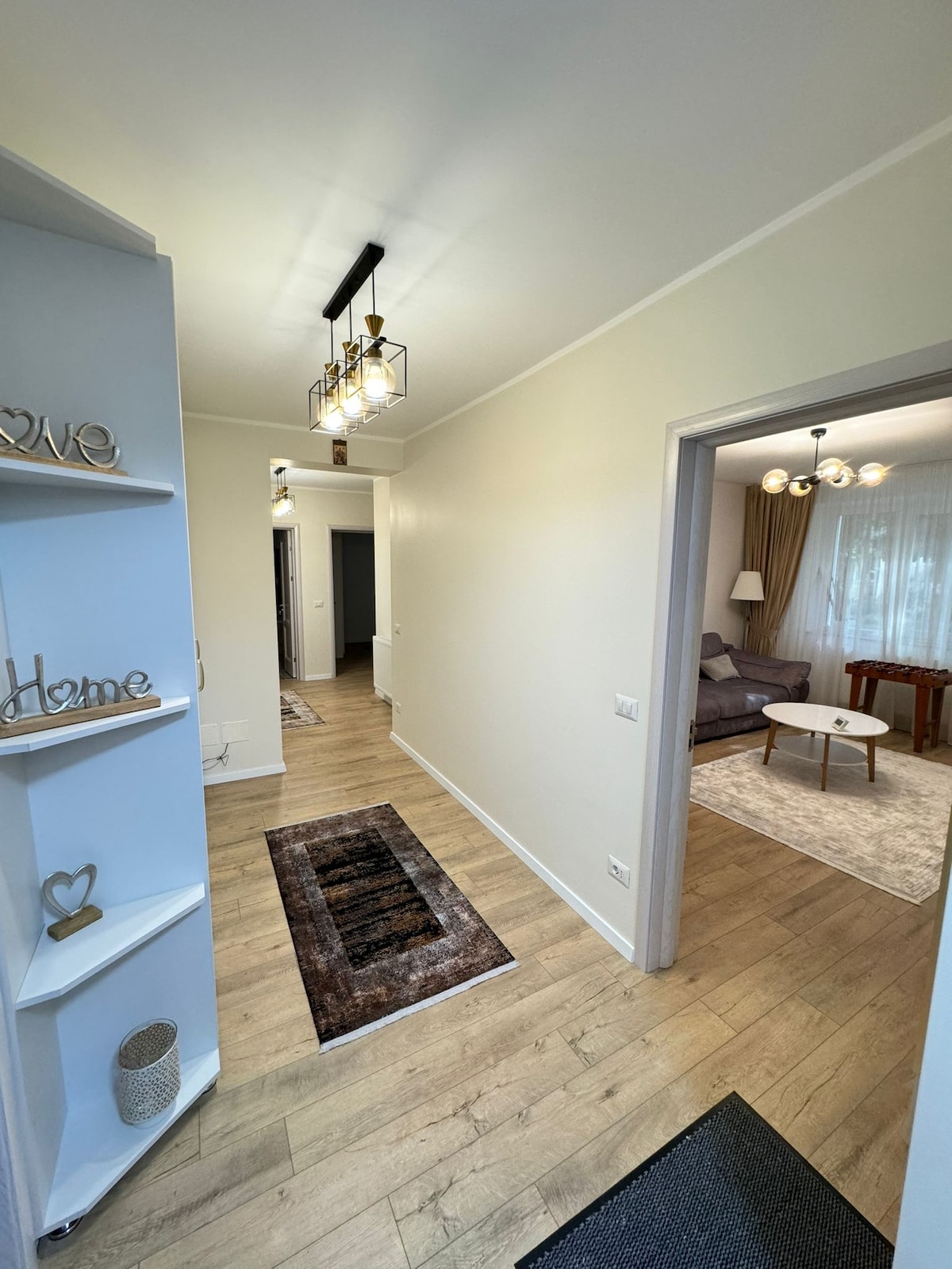 A well-lit hallway features warm wood flooring and neutral walls. A contemporary light fixture hangs from the ceiling. To one side, a cozy living area is visible, showcasing a gray sofa and a small table. A runner rug adds texture to the space.