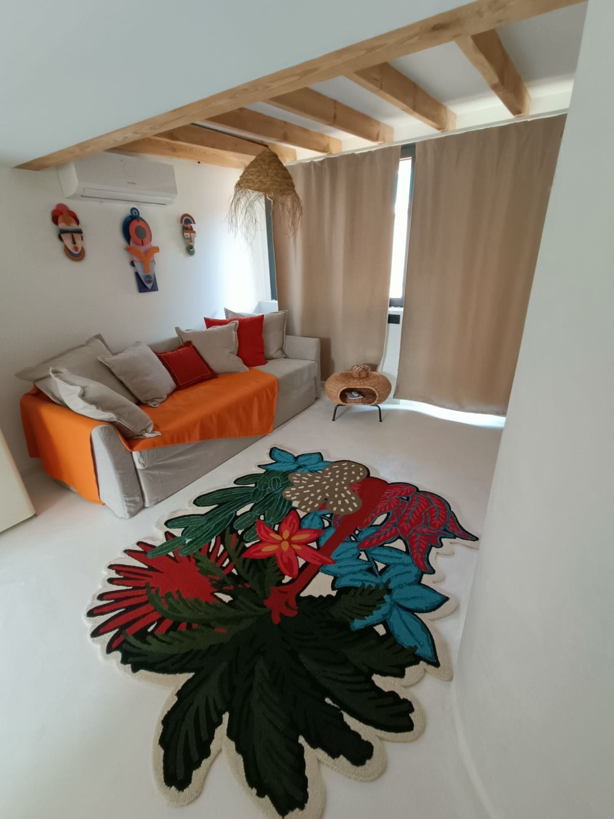 A cozy sitting area is furnished with a gray sofa adorned with colorful cushions in red and orange. A large, intricately patterned area rug featuring vibrant floral designs anchors the space. Soft curtains provide privacy while light filters in from a nearby window.