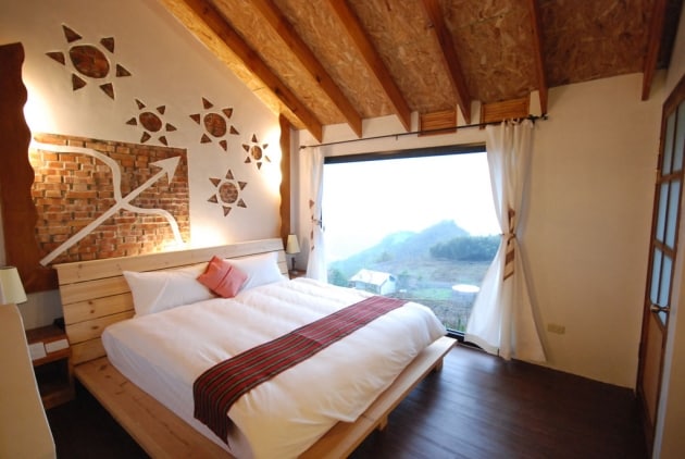 A spacious sleeping area features a large bed with a light blanket and decorative pillows, set against a backdrop of artistic wall elements. A wide window allows natural light to fill the room, offering a view of the surrounding mountainous landscape.