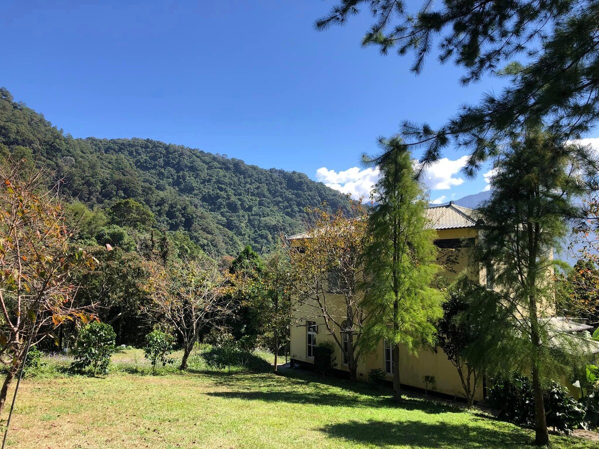 A serene outdoor area is depicted, featuring a lush green lawn bordered by trees. A gently sloping mountain landscape is visible in the background under a clear blue sky, emphasizing the peaceful natural surroundings.
