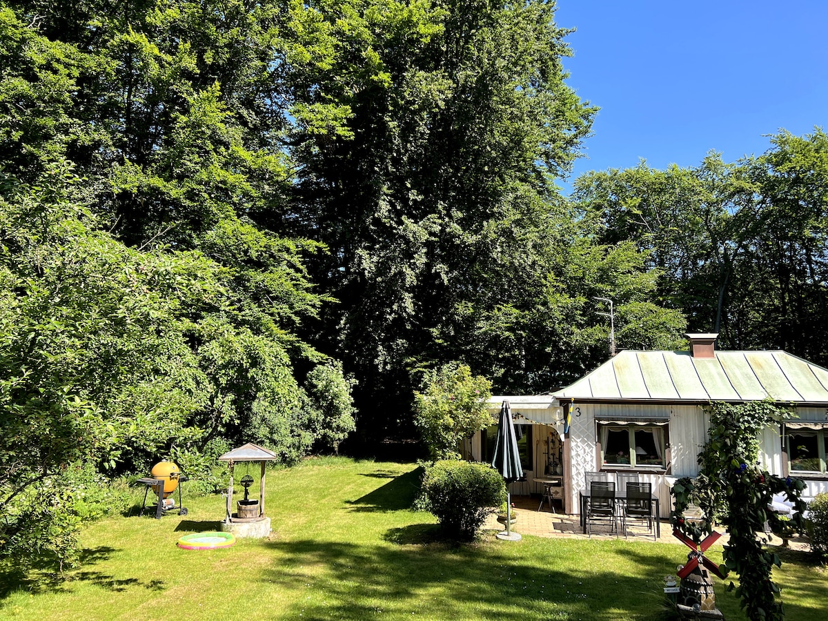 A charming cottage is set against a backdrop of lush greenery. The spacious garden features a gazebo, a grill area, and vibrant plant life. Sunlight filters through the trees, creating a serene environment ideal for outdoor relaxation.