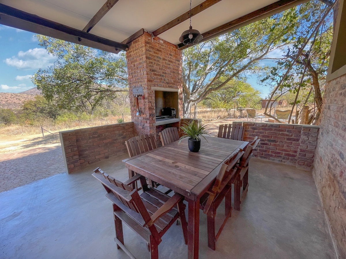 An outdoor dining area is featured with a wooden table surrounded by matching chairs. A brick-built fireplace provides a space for grilling. The area is framed by trees, with open views of the natural surroundings, creating a relaxed ambiance.