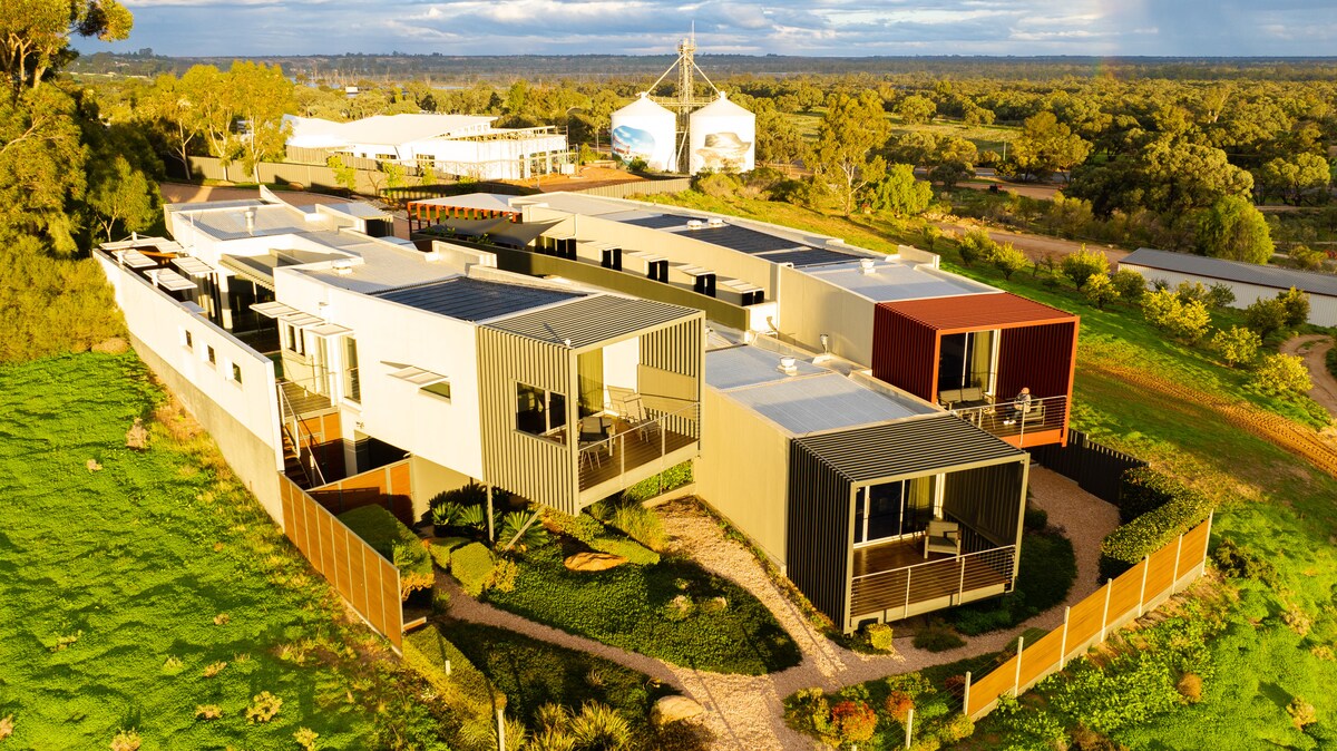 An aerial view of a contemporary retreat is displayed, featuring several interconnected structures in neutral tones. Surrounding green landscapes are visible, along with pathways leading to outdoor seating areas. Sunlight highlights the building's modern design, creating a harmonious blend with the natural environment.