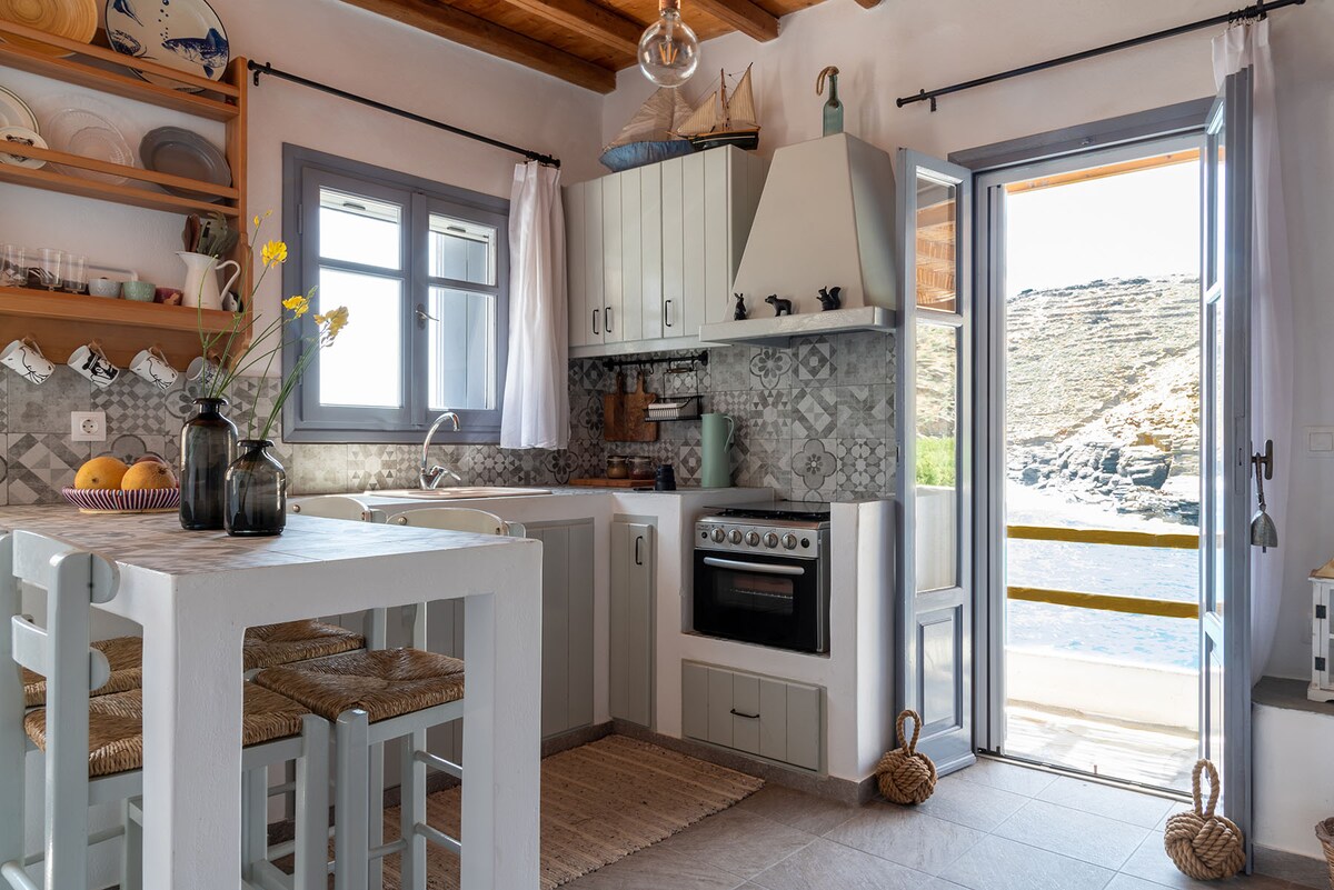 A well-equipped kitchen features light cabinetry and a stylish backsplash. Natural light floods the space through the open window and door, which offer views of the sea. A simple dining table with light-colored chairs complements the inviting atmosphere.