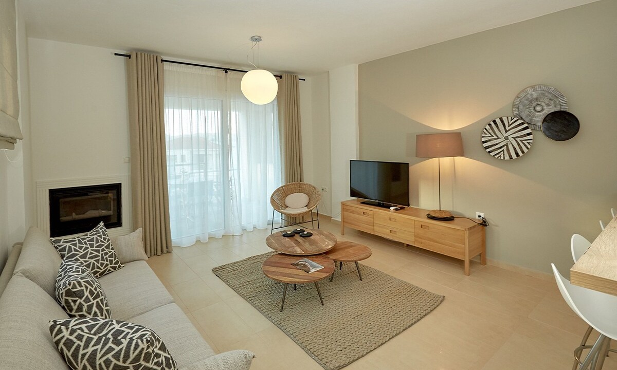 The living area features a neutral color palette with natural light filtered through sheer curtains. A comfortable sofa with decorative cushions is positioned near a wooden coffee table. A modern entertainment unit displays a television, and a stylish floor lamp adds warmth to the space.