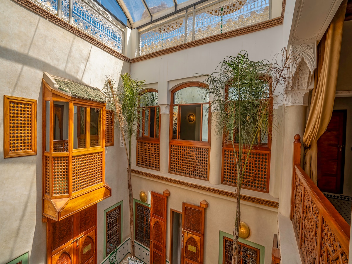 An airy interior courtyard is surrounded by wooden lattice windows and doors, allowing natural light to filter through. Two tall palm trees add a touch of greenery. Intricate archways and decorative elements highlight the craftsmanship typical of Moroccan architecture.