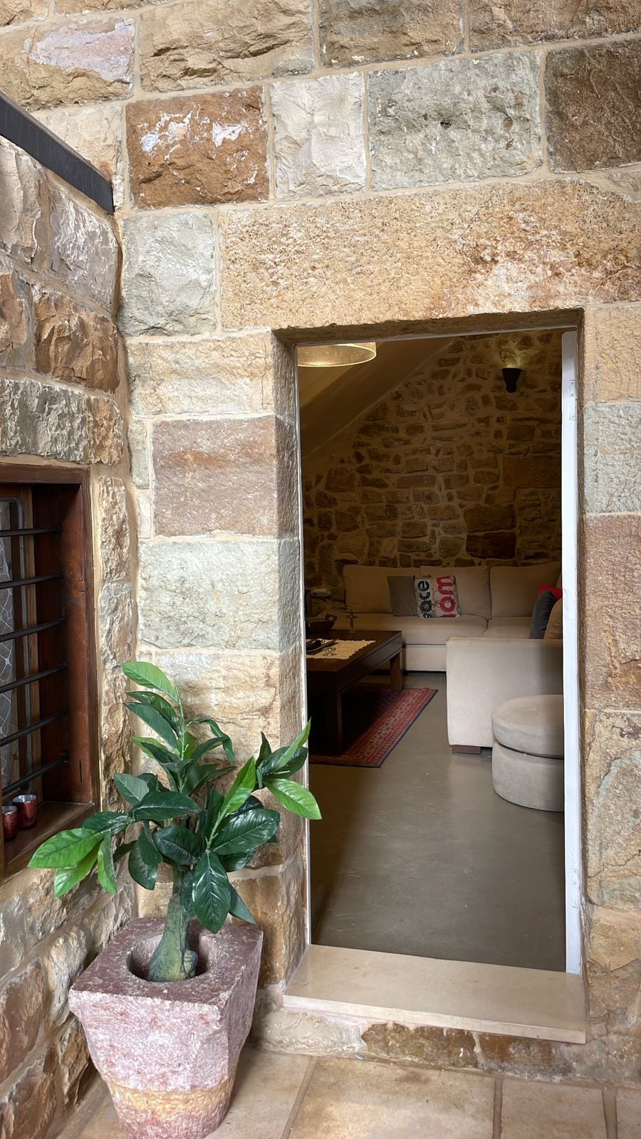 An entrance framed by textured stone walls leads into a cozy living area. A potted plant adds greenery near the doorway, while soft furniture can be glimpsed in the inviting space beyond. Natural light illuminates the interior, enhancing the warm tones of the stone.