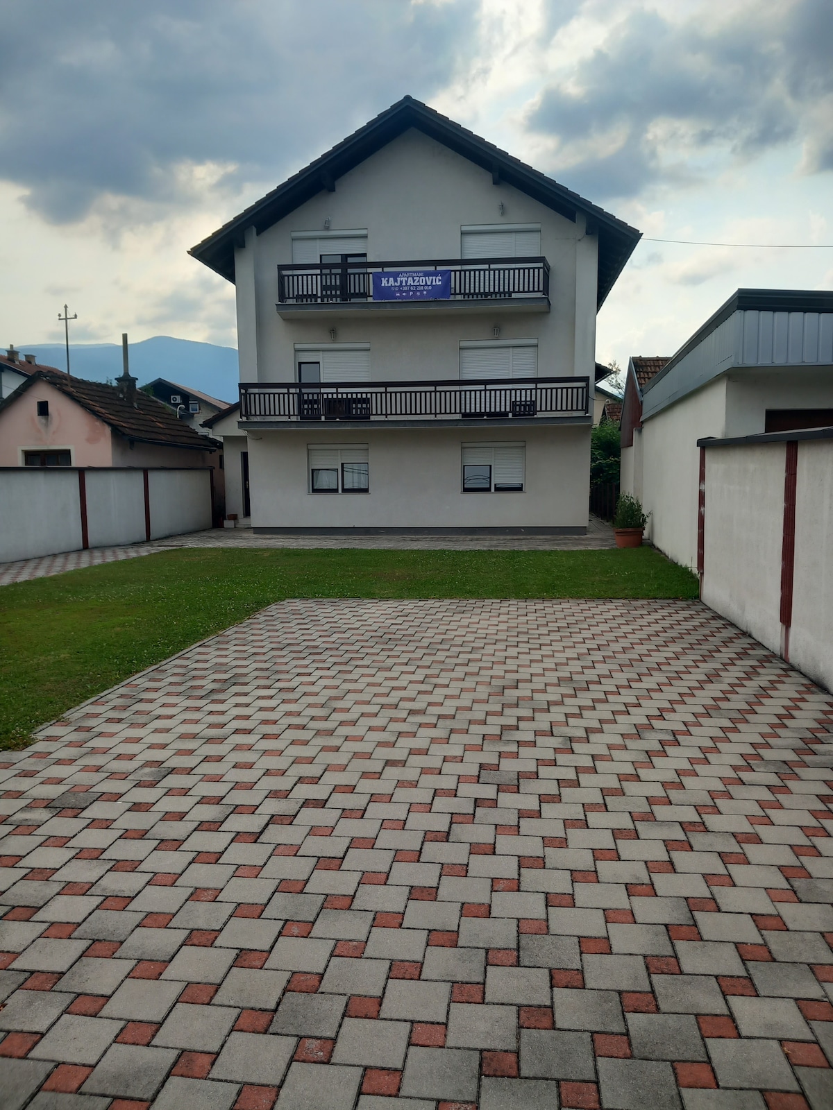 A three-story building is shown, featuring a flat roof and a balcony on the upper floor. The surrounding area is paved with patterned bricks, and a small grassy lawn is visible in front. Nearby structures are partially obscured by a low wall.