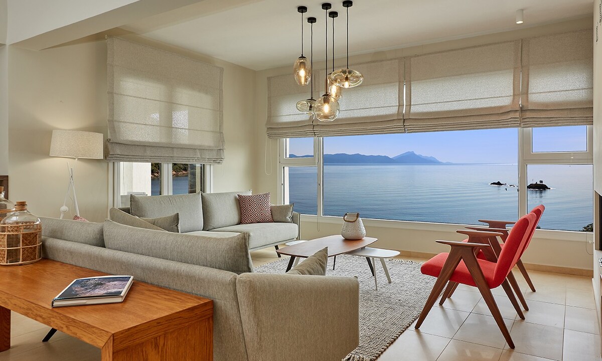 A spacious living area features large windows that provide panoramic sea views. Comfortable seating, including a grey sofa and a red accent chair, is arranged around a wooden coffee table. Soft lighting is created by stylish pendant fixtures, adding warmth to the neutral decor.