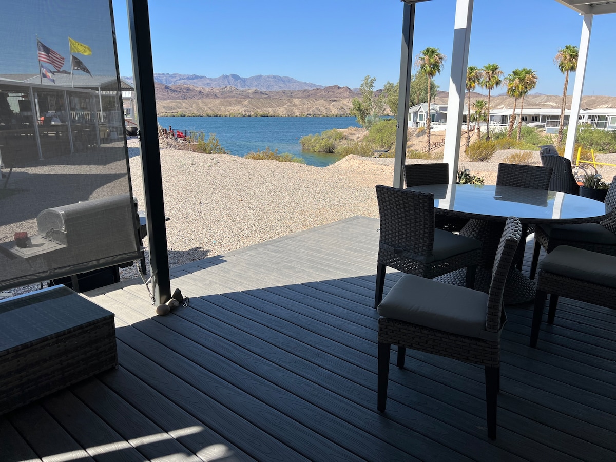 A covered deck offers a view of the lake, with several palm trees visible in the background. Outdoor seating, including a round table and four chairs, is arranged on the wooden floor, providing a comfortable space to relax and enjoy the surrounding natural landscape.