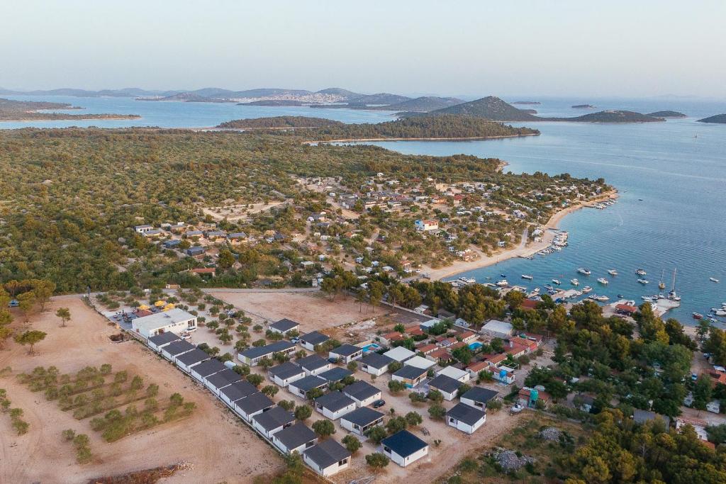 An aerial view of a coastal area reveals a mix of natural greenery and developed spaces. Small boats rest in a serene bay, with a collection of neatly arranged accommodations visible along the shoreline. The presence of distant islands adds to the tranquil landscape.