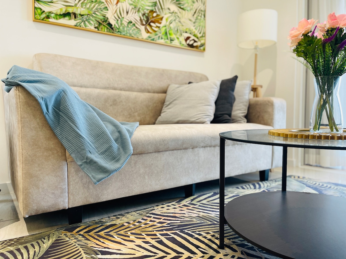 A contemporary three-seater sofa is positioned in a living area, adorned with various cushions in neutral tones. An elegantly designed coffee table sits in front, complemented by a vase of fresh flowers. A patterned area rug enhances the overall aesthetic, while a lamp provides soft lighting.