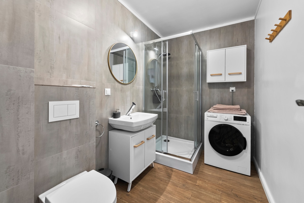 An elegantly designed bathroom features a walk-in shower enclosed by clear glass. A round mirror above a white vanity reflects the light, complemented by modern cabinetry. A neatly stacked set of towels is placed on the vanity, while a washing machine is situated beside it.