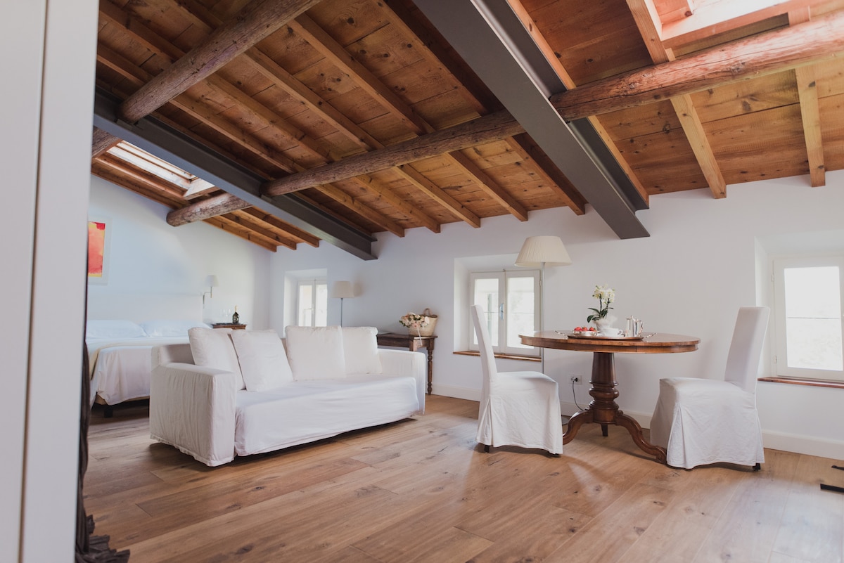 An open living area features a white sofa and a circular dining table. Sunlight enters through several windows, highlighting the wooden beams and flooring. A spacious bed is positioned in the background, contributing to the airy and inviting atmosphere.